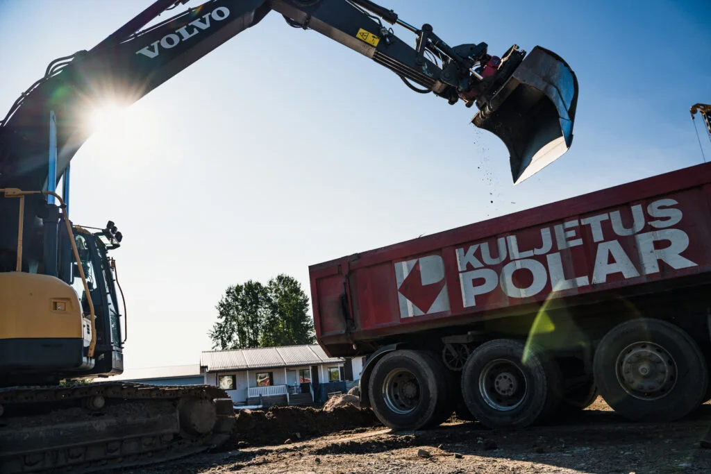 Talon pohjatyöt näkyvät loppukesän kuvassa: keltainen kaivinkone lastaa poisvietävää maa-ainesta punalavaiseen kuorma-autoon.