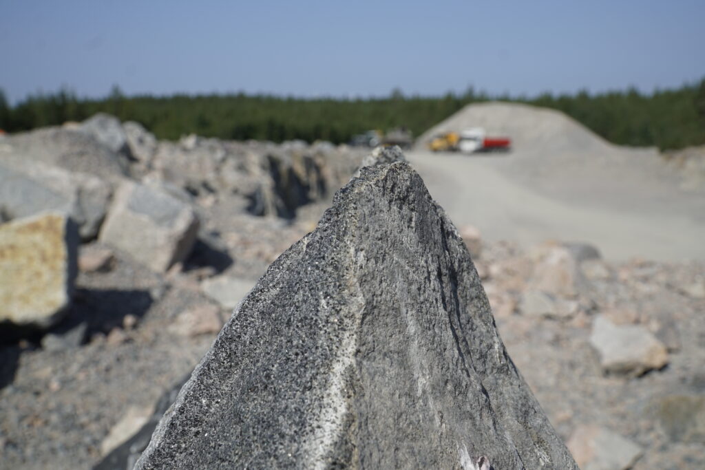 Tukimuurit: lähikuva kiviaineksesta, jossa terävä kärki: näyttää vuorelta. Taustalla maa-aineksen ottopaikka ja murskekasa sekä työajoneuvoja.
