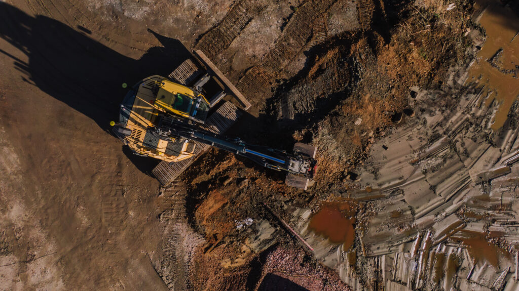 Ilmakuva noin 50 metristä, kamera on kuvannut suoraan alaspäin. Keltainen kaivinkone urakoimassa rakennustontilla.